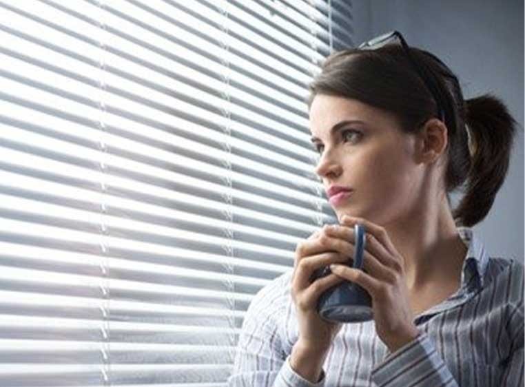 Woman holding a coffee mug looking out of a window with blinds.